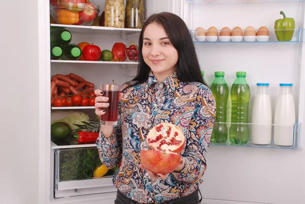 Mooi lachende jonge meisje houdt een glas granaatappelsap en granaat op de achtergrond van de koelkast — Stockfoto