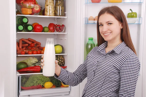Молодая девушка держит бутылку молока, стоя возле открытого холодильника — стоковое фото