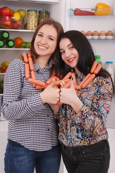 Two young girls holds sausages on the refrigerator background — Stock Photo, Image