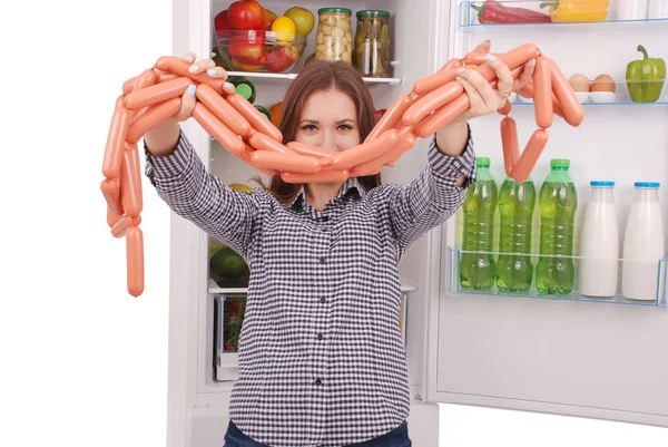 Молодая девушка держит сосиски на заднем плане холодильника — стоковое фото
