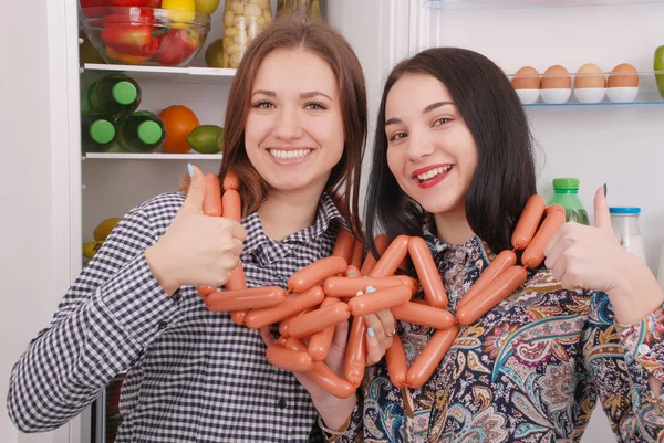 Две молодые девушки держат сосиски на заднем плане холодильника — стоковое фото