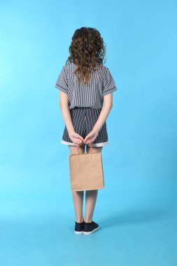 Mavi arka planda izole edilmiş bir paketolan çocuk. Tatil hediyesi, alışveriş. Alışveriş merkezinde çocuk mutlu alışveriş. Uzun kıvırcık saçlı mutlu bir kız. Yüksek çözünürlüklü fotoğraf. Tam alan derinliği.