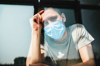 Virüs yüzünden karantinada olan bir adam maske takıp güneşli bir günde pencereden dışarı bakar.