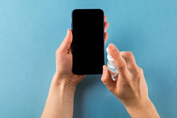 Woman Wiping Mobile Phone Screen With Sanitizer on blue background. Cleaning smart phone screen to prevent corona virus Covid 19