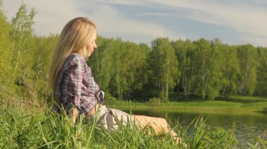 Yeşil çimenlerin üzerinde oturan göl kıyısı tarafından kadın rahatlatır. Yaz tatil kavramı.