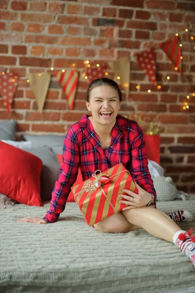 Mujer joven se sienta en una cama y sostiene el regalo de Navidad en la mano . — Foto de Stock