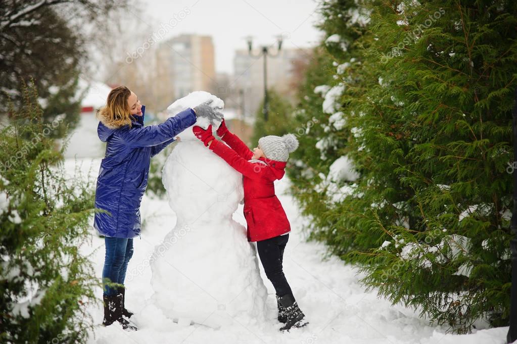 Mother with daughter of younger school age build a snowman in the park.