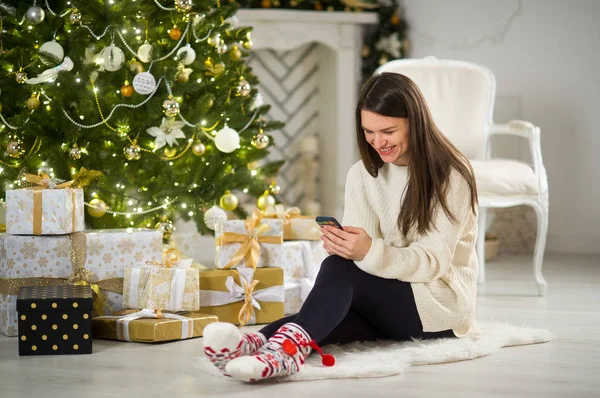 Niza morena chica se sienta cerca del árbol de Navidad con el teléfono móvil en las manos . — Foto de Stock