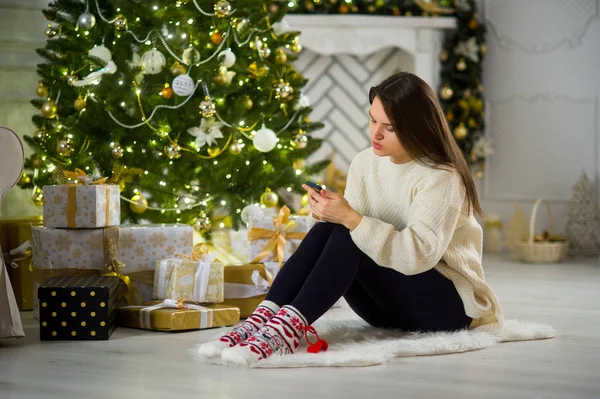 Söt mörkhårig flicka sitter nära julgran med mobiltelefonen i händer. — Stockfoto