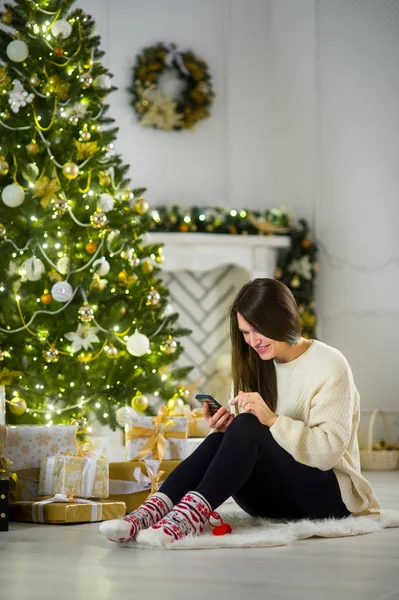 Linda chica de pelo oscuro se sienta cerca del árbol de Navidad con el teléfono móvil en las manos . — Foto de Stock