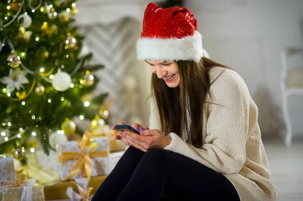 Bella ragazza in berretto di Babbo Natale si siede vicino all'albero di Natale con il telefono cellulare in mano . — Foto Stock