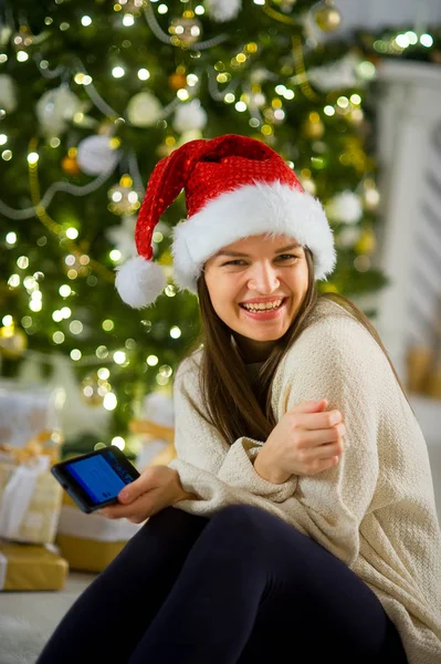 Bella ragazza in berretto di Babbo Natale si siede vicino all'albero di Natale con il telefono cellulare in mano . — Foto Stock