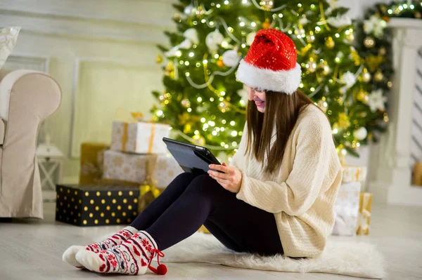 Charmig tjej i tomtens cap sitter närliggande en julgran med laptop. — Stockfoto