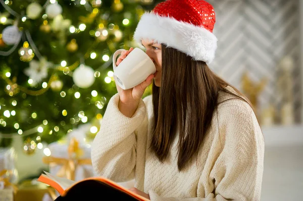 Chica en la gorra de Santa bebe café cerca del árbol de Navidad . — Foto de Stock