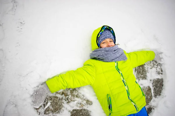 Der fröhliche Junge liegt auf Schnee und macht Engelsflügel. — Stockfoto