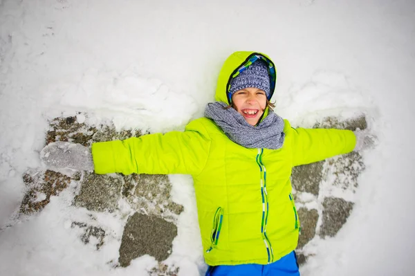 Der fröhliche Junge liegt auf Schnee und macht Engelsflügel. — Stockfoto
