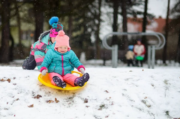 Two sisters in bright ski suits ride the sledge in winter day. — Stock Photo, Image