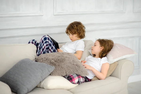 Zwei Kinder, der Junge und das Mädchen, liegen mit dem Tablet in der Hand auf einem Sofa. — Stockfoto