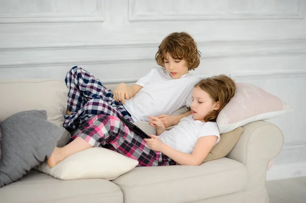 Dos niños, el niño y la niña, yacen en un sofá con la tableta en las manos . — Foto de Stock