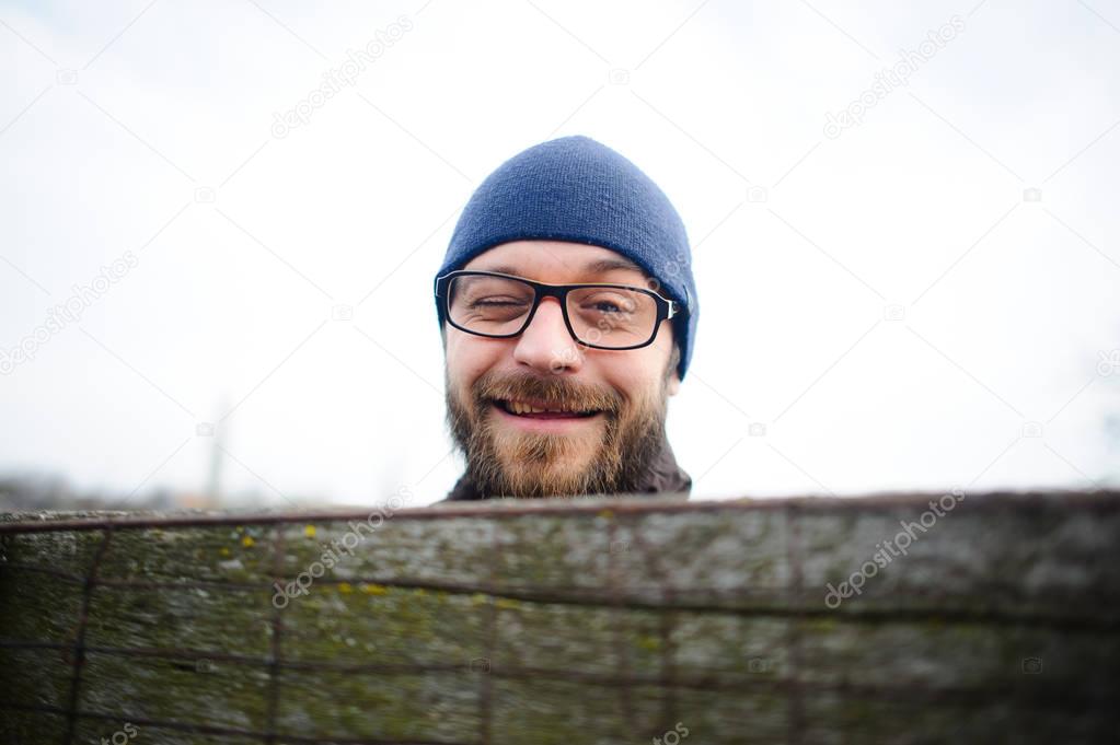 Funny young man with glasses and a beard looks out from behind the fence. He