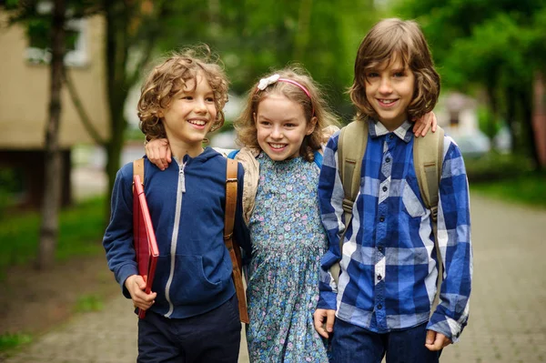 Tre små skolelever, två pojkar och en flicka, stå i en omfamning på skolgården. — Stockfoto