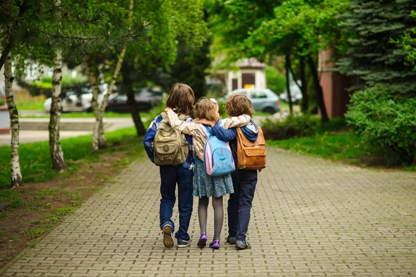 Tres pequeños estudiantes de la escuela, dos chicos y la chica, van en un abrazo a la escuela —  Fotos de Stock