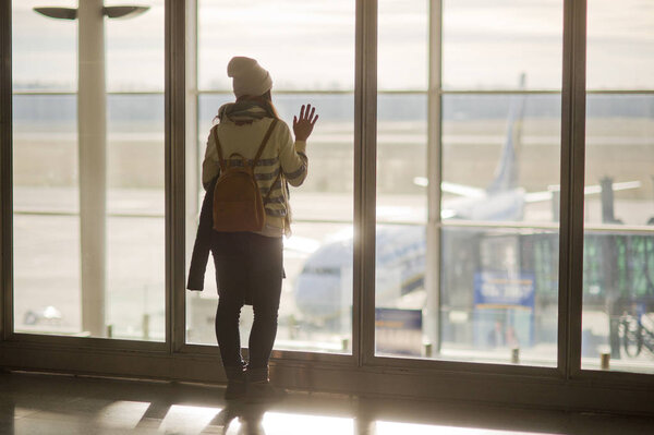 A woman with a backpack standing at the large window in the airport.