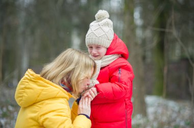 Young mother consoles the crying child. The girl has fallen during walk. For her it is sick and offensive. The loving mother calms the daughter. Against the background of the winter park. clipart