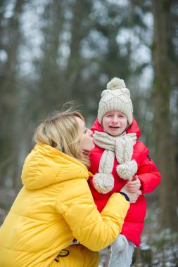 Young mother consoles the crying child. The girl has fallen during walk. For her it is sick and offensive. The loving mother calms the daughter. Against the background of the winter park. clipart