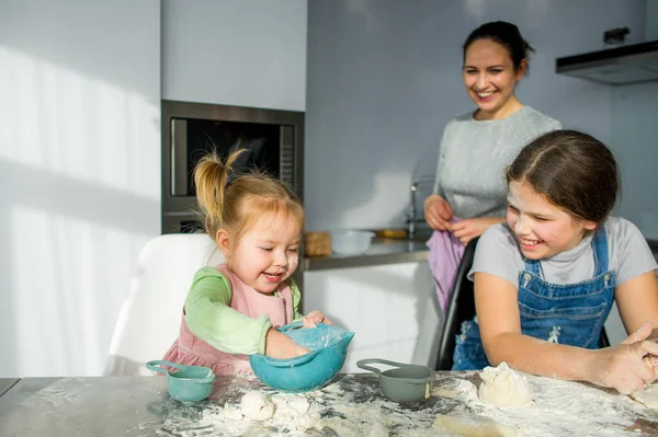 Mom teaches two little daughters to cook from dough. Children with pleasure are engaged in kitchen work.