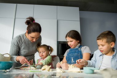 Anne üç çocuğuna yemek yapmayı öğretiyor. Aile hamurdan bir şeyler hazırlıyor. Mutfak masasında gerekli ürünler var. Faydalı ve eğlenceli bir eğlence..