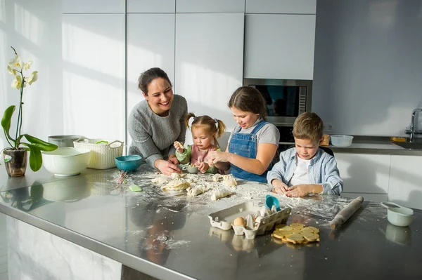 Mother teaches her three children to cook. Family is preparing something from the dough. There are necessary products on the kitchen table. Useful and fun pastime.