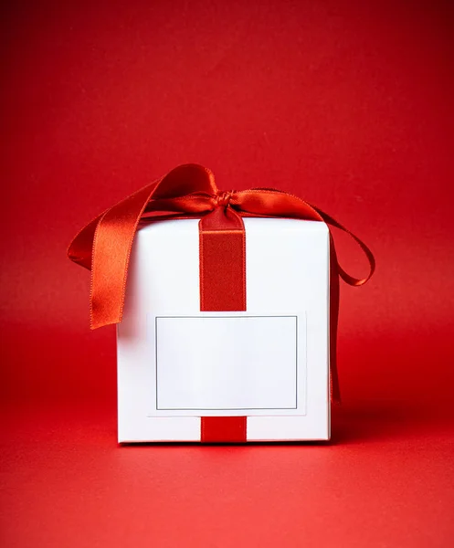 Weiße Geschenkschachtel mit leerem Lable und rotem Satinband isoliert auf rotem Studiohintergrund — Stockfoto