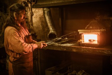 Senior blacksmith heats item before forgingin smithy clipart