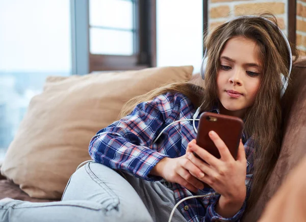 Menina bonita em fones de ouvido usando smartphone em um sofá — Fotografia de Stock