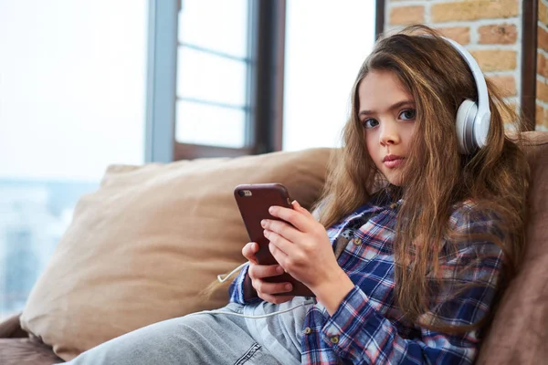 Hermosa niña en auriculares con teléfono inteligente en un sofá —  Fotos de Stock