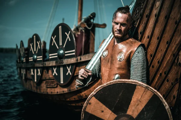 Poważne viking wojownika z mieczem i tarczą stojący w pobliżu Drakkar nad brzegiem morza. — Zdjęcie stockowe
