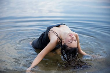 Kumsalda ıslak saçlı bir kadın oturuyor. Siyah bir elbise giyiyor..