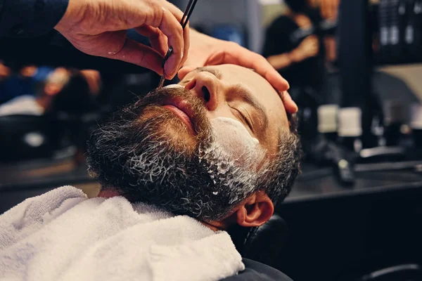 Barber shaving bearded male with a sharp razor — Stock Photo, Image