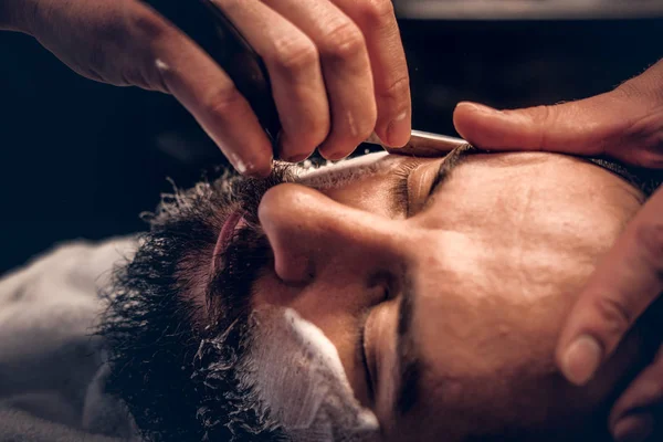 Barber shaving bearded male with a sharp razor — Stock Photo, Image
