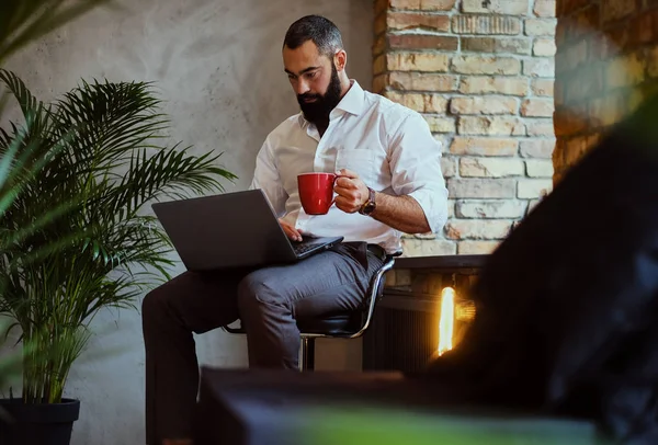 Hombre barbudo urbano bebe café y usa un portátil . — Foto de Stock