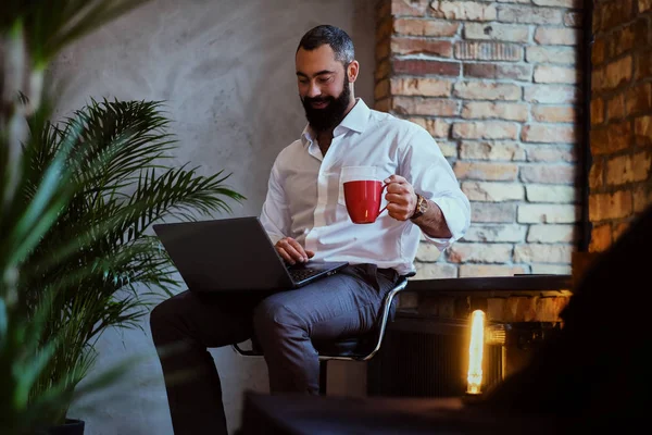 Városi szakállas férfi italok kávé és használ egy laptop. — Stock Fotó