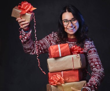 Gülümseyen bir esmer kadın kırmızı bir Kazak giymiş ve gözlük tutan Noel hediyeleri gri arka plan üzerinde.