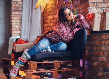 Esmer kadın gözlük tam vücut görüntü bir kot pantolon ve ahşap bir kanepe Noel dekorasyonu ile bir odada poz kırmızı Kazak giymiş.