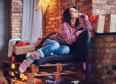 Esmer kadın gözlük tam vücut görüntü bir kot pantolon ve ahşap bir kanepe Noel dekorasyonu ile bir odada poz kırmızı Kazak giymiş.