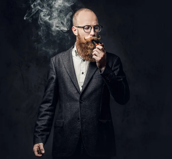Kızıl saçlı sakallı erkek takım elbise ve gelenek pipo koyu gri arka plan üzerinde gözlük giyinmiş.