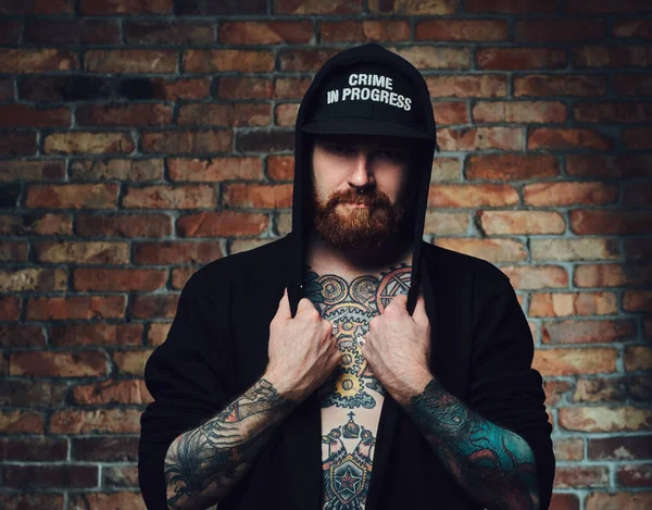 Kızıl saçlı sakallı erkek silah ve göğsünde, dövmeli portresi giymiş siyah spor ceketli bir tuğla duvarın üzerinden.