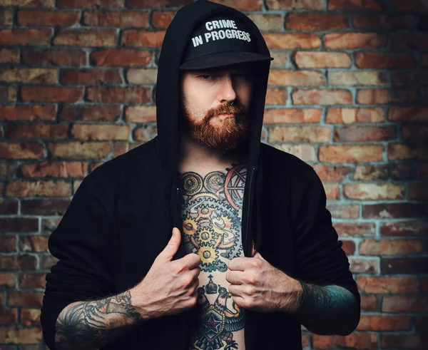 Portrait of redhead bearded male with tattoos on arms and chest, dressed in black sports jacket over the wall of a brick.