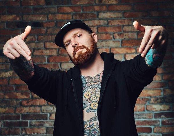 Portrait of redhead bearded male with tattoos on arms and chest, dressed in black sports jacket over the wall of a brick.