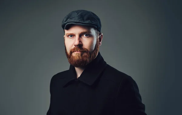 Portrait of Redbeard elegant male dressed in a tweed flat cap and a black jacket over grey background.